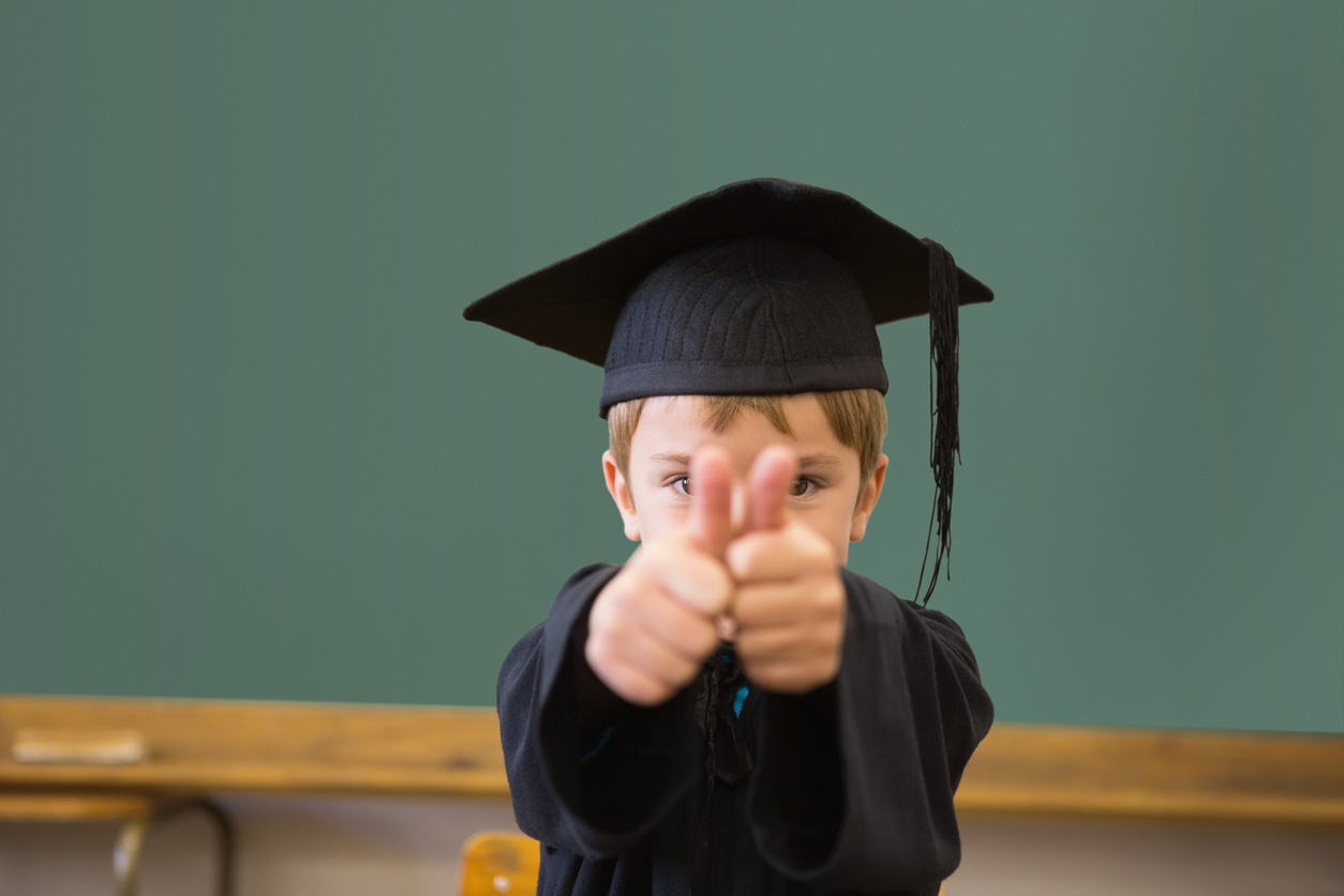 Cute pupil in graduation robe smiling at camera in classroom at the elementary school