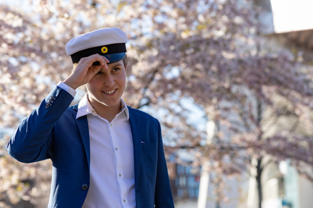 A teenage boy in a suit and a Swedish graduation hat. He is standing under cherry blossom trees.