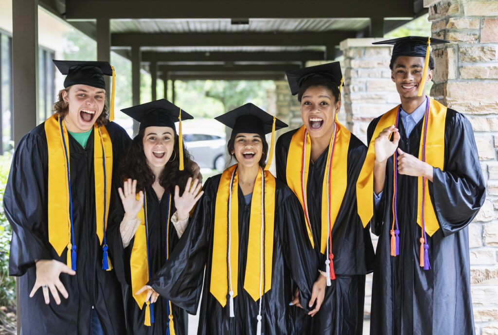 A group of five multiracial high school graduates standing together outside the school, wearing their caps and gowns, waving and shouting, looking at the camera.