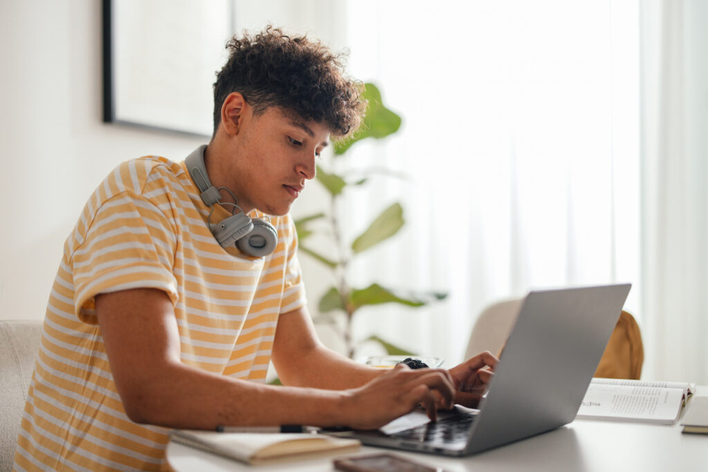 Handsome teenage boy sitting at the table at home doing his homework or studying. He is looking serious and focused while using his laptop computer and typing on the keyboard. He is wearing Bluetooth headphones around his neck.