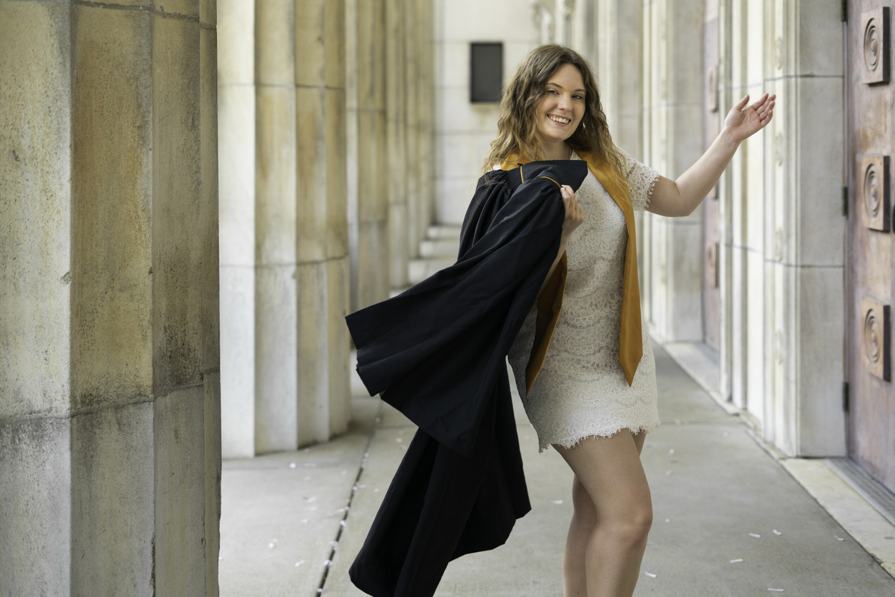 Blond female wearing a with a gold sash over a white dress holds her black graduation gown and mortarboard cap in her hand while standing outdoors after college graduation