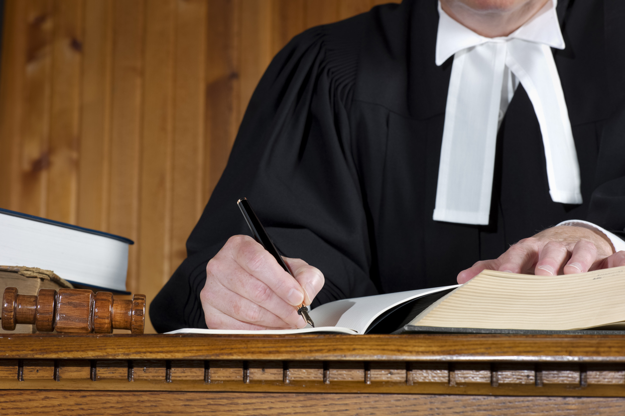 A judge takes notes at a desk or bench during a trial. Robes are traditional British Commonwealth Barrister gowns.