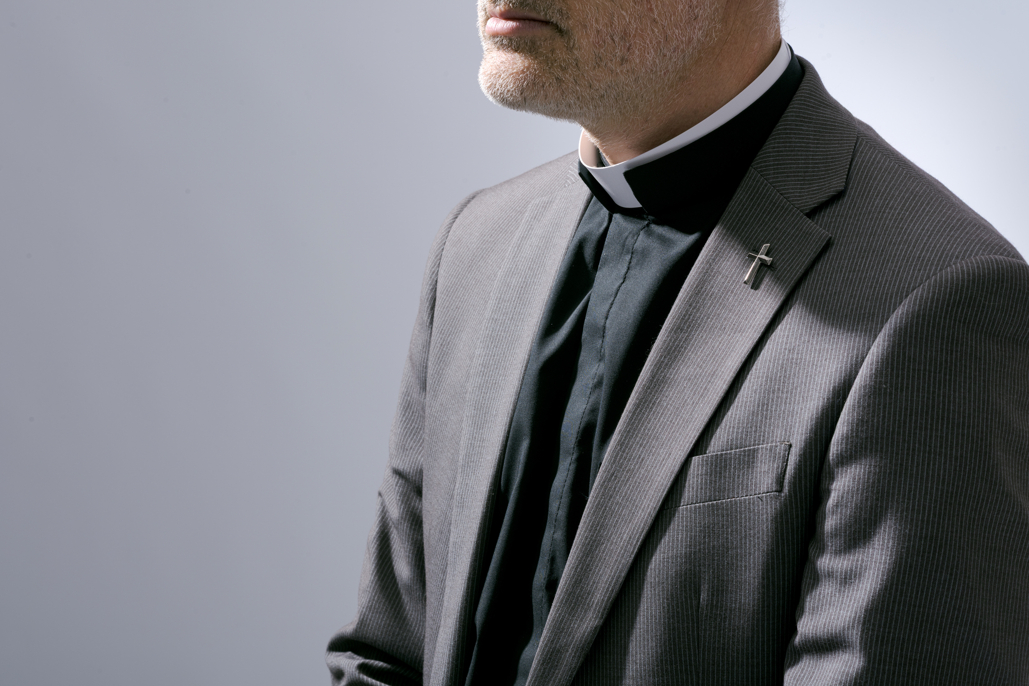 Catholic parish priest, half-length on gray background. Fancy dress and white clerical collar. A silver groce stands out on the lapel of his jacket, a symbol of his faith.