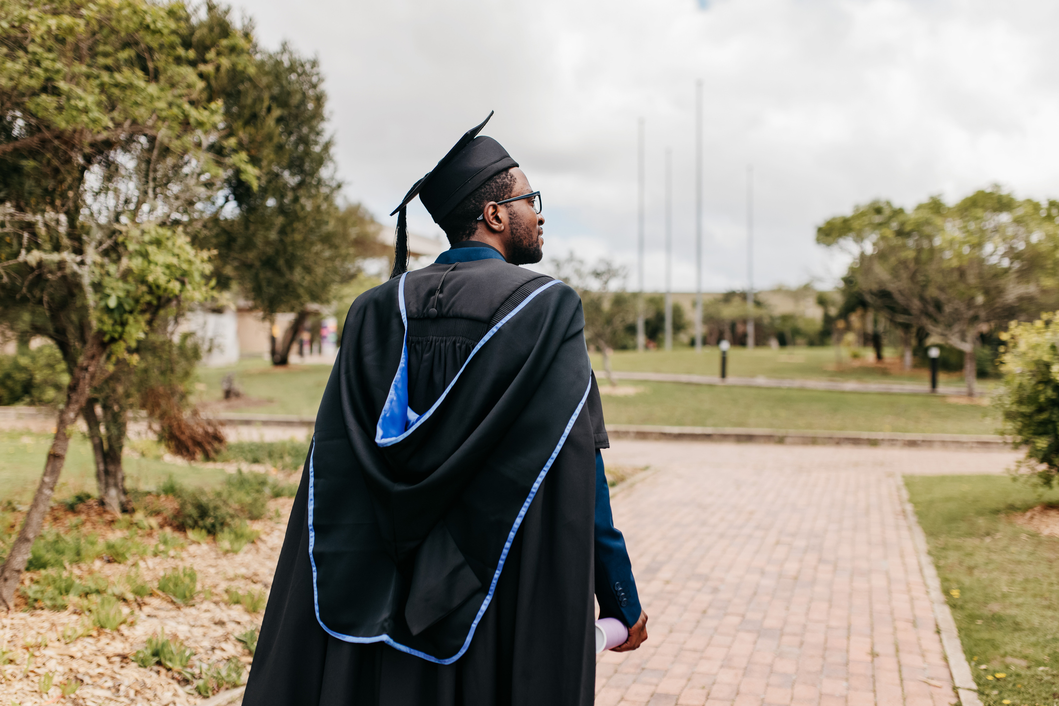 Young male graduate with beard wearing graduation gown and mortarboard walking and looking sideways into distance on campus grounds